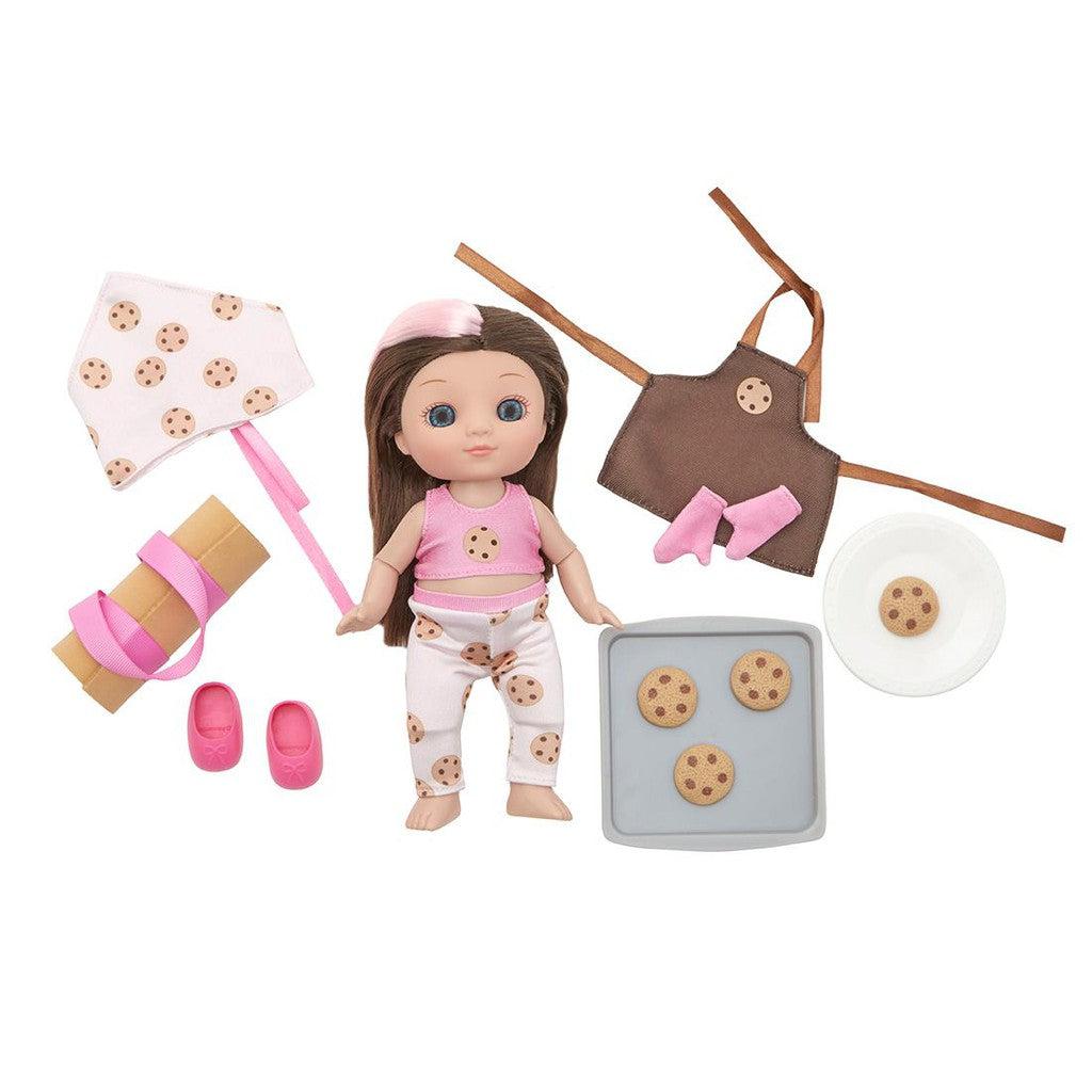 A posable baker doll with long brown hair, surrounded by baking accessories like a rolling pin, baking sheet, brown apron, pink shoes, and cookie-themed clothing.