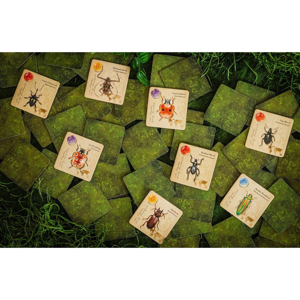 Cards featuring diverse beetle families and game tokens are scattered on a table draped in green fabric and foliage, resembling a jewel box of nature's treasures.