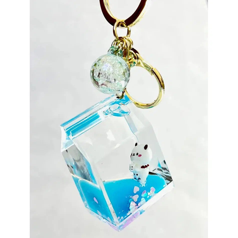 A keychain featuring a clear, cube-shaped acrylic charm with a miniature white bear playfully floating on blue liquid. This delightful key charm, part of our assorted designs collection, is completed with a decorative glass bead and gold clasp.