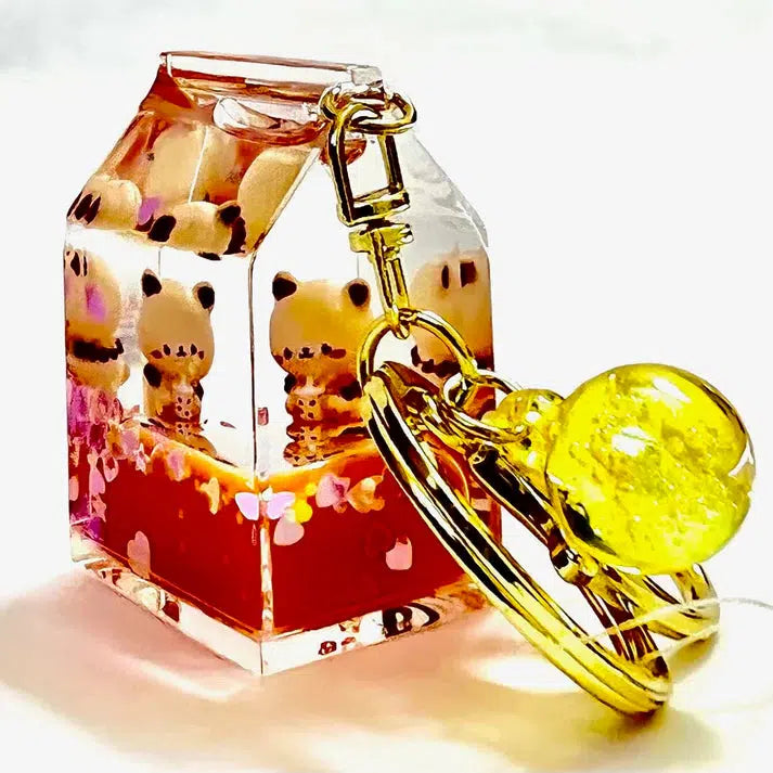 Keychain with a small transparent kitty milk carton charm, featuring silly cat figures inside and a yellow orb attached to gold rings.