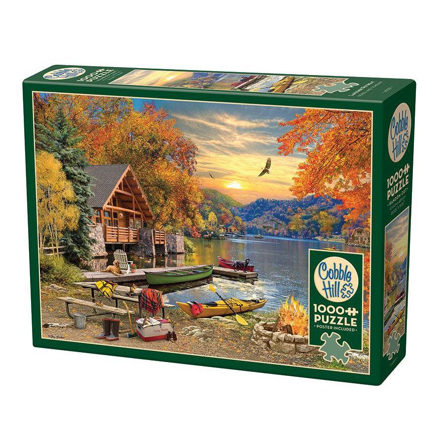 1000 pc puzzle of a cabin next to a lake in a forest. The sun is setting and the trees are orange. There are kayaks on the shore