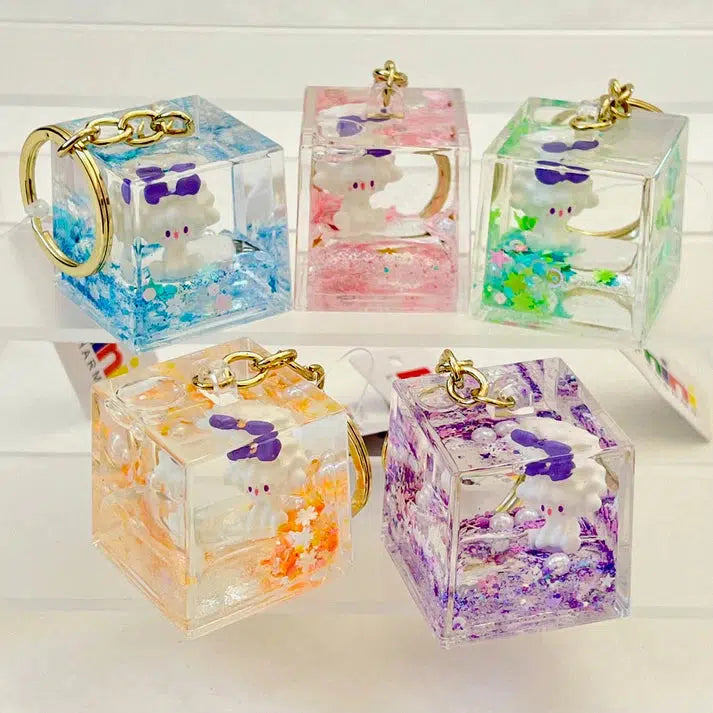 Five cube-shaped keychains feature colorful liquid and a small white character inside each, showcasing silly animals in an array of vibrant colors: blue, pink, green, orange, and purple. With assorted designs, these lamb floaty key charms add a playful touch to any accessory collection.