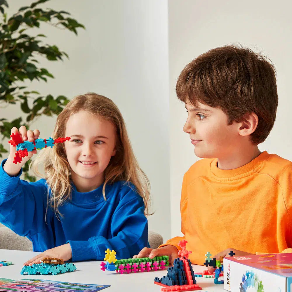 Two children sit at a table, immersed in creativity as they build colorful structures with Plus-Plus blocks. The girl enthusiastically presents her creation, while the boy observes with curiosity. A plant quietly thrives in the background, adding to their little Colorverse of imagination.