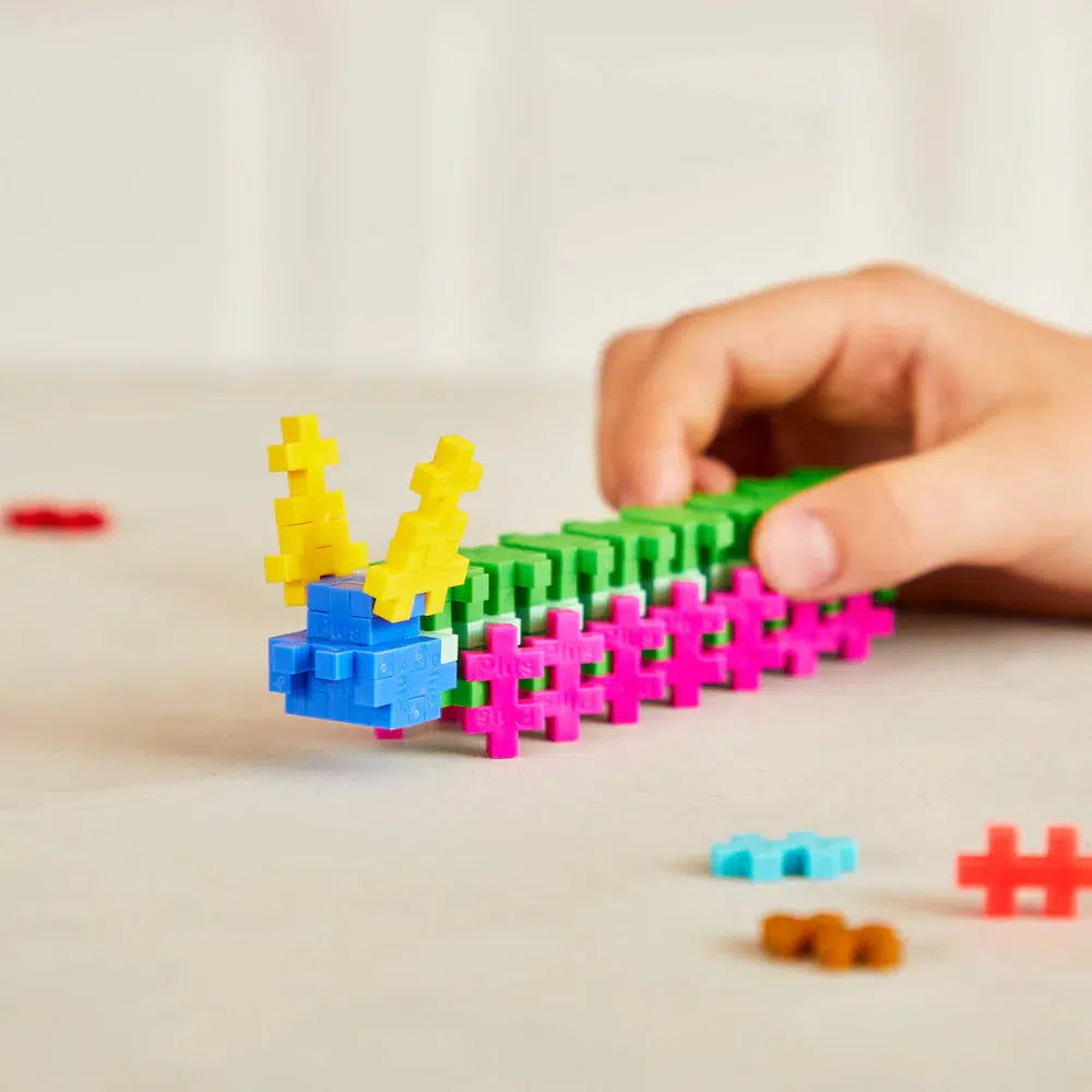 A hand is showcasing creativity by assembling a vibrant caterpillar-like structure with Plus-Plus interlocking plastic blocks on a table, creating a mini Colorverse in the process.