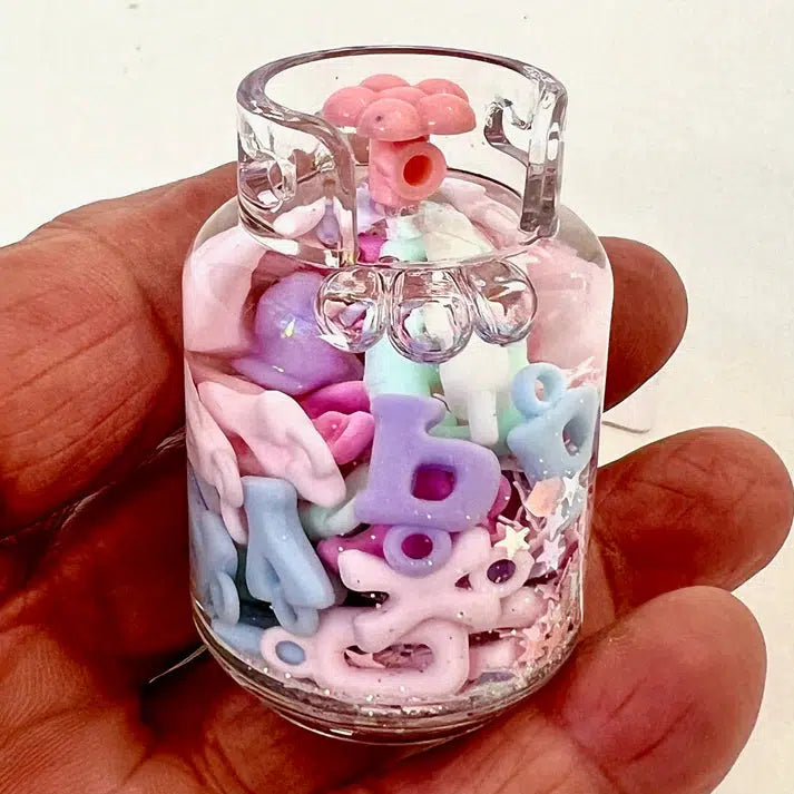 A hand holds a small glass jar brimming with colorful pastel polymer clay letters and shapes, alongside an array of assorted designs.