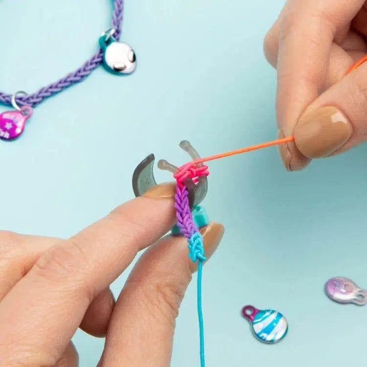 Close-up of hands using a tool to weave colorful yarn, with a blue background and small charms scattered around.