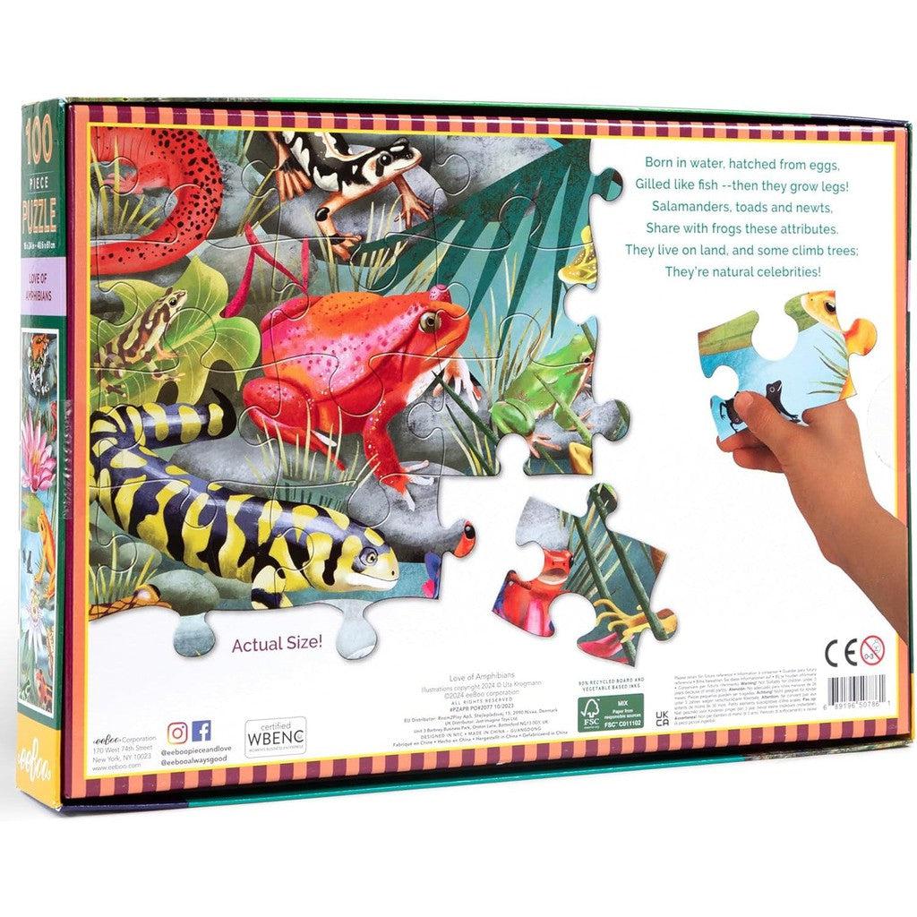 The eeBoo Kids Jigsaw Puzzle is a colorful 100-piece set featuring amphibians like frogs and salamanders. A hand holds a puzzle piece as text highlights the love of amphibian traits and their habitats.