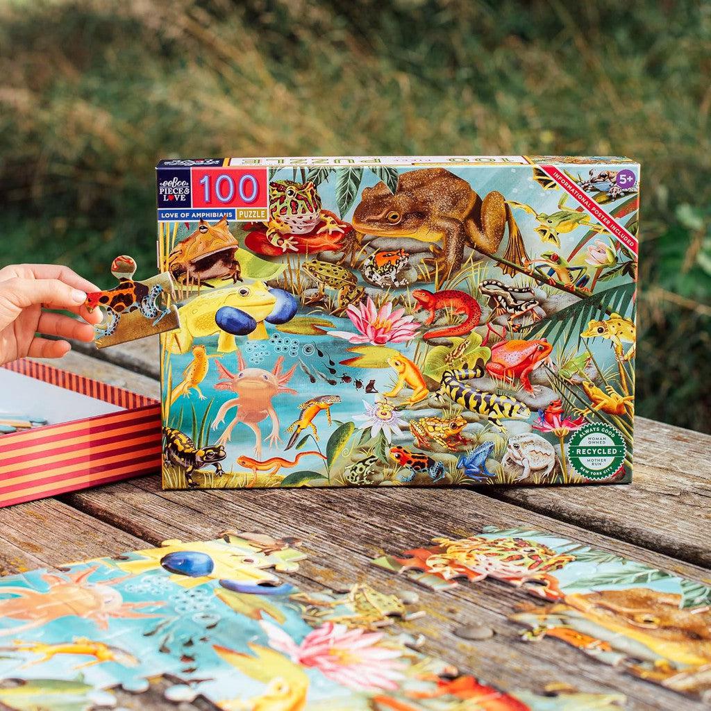 A person assembles an eeBoo 100-piece jigsaw puzzle featuring a colorful variety of animals, including frogs and fish, on a wooden table outdoors.