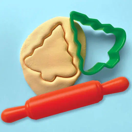 A green tree-shaped cookie cutter and a red rolling pin rest on a light blue background, with a festive activity underway: a tree shape delicately cut out of yellow dough, evoking the magic of cookie-shaped ornaments.