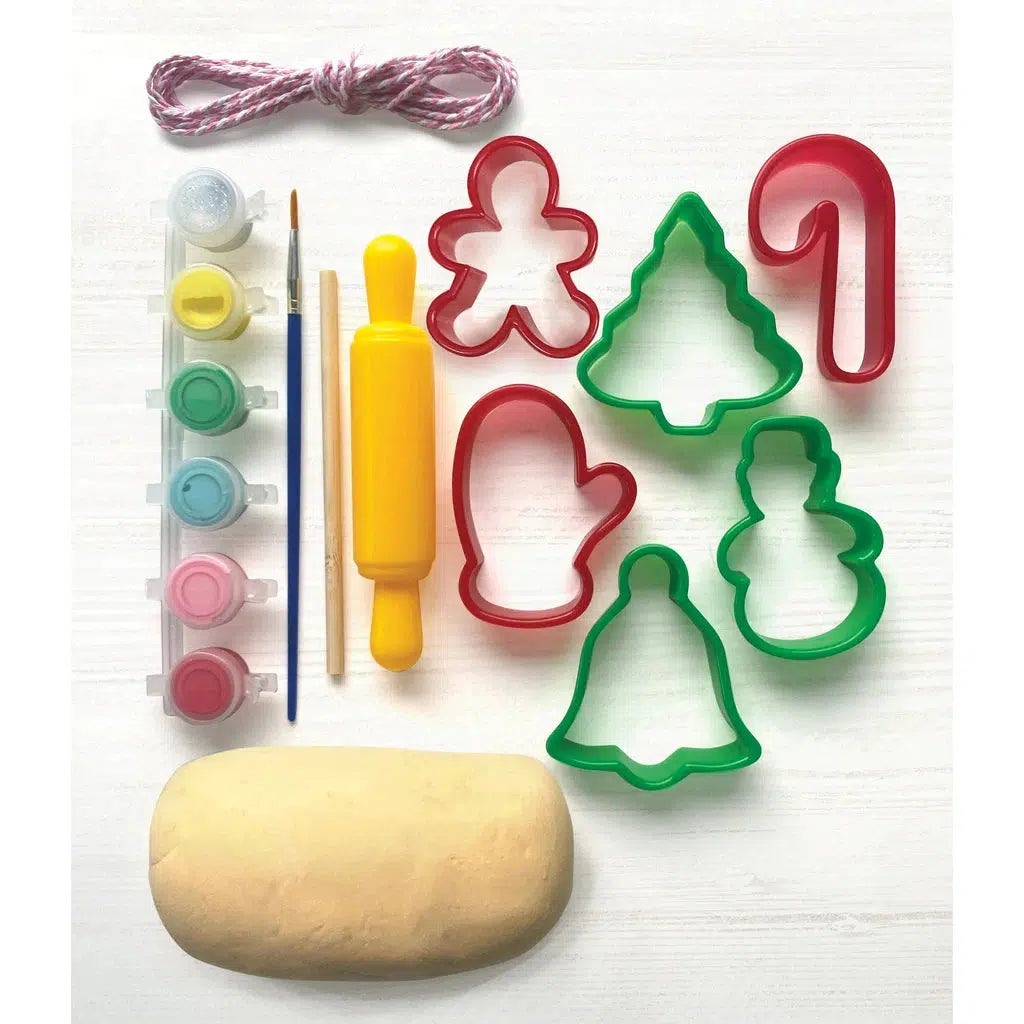 Dough, a small rolling pin, cookie cutters, paints, and brushes are arranged on a white surface. Cookie cutters are in shapes like a gingerbread person, tree, mitten, bell, and candy cane—perfect for creating air-dry clay ornaments as a festive activity.