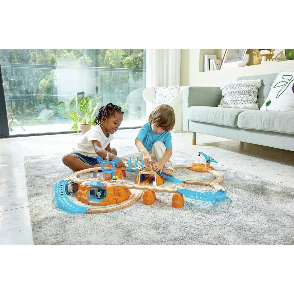 Two children play with a train set on a living room rug, their eyes filled with railway excitement. Large windows reveal a garden outside, while a cozy sofa with pillows sits in the background.