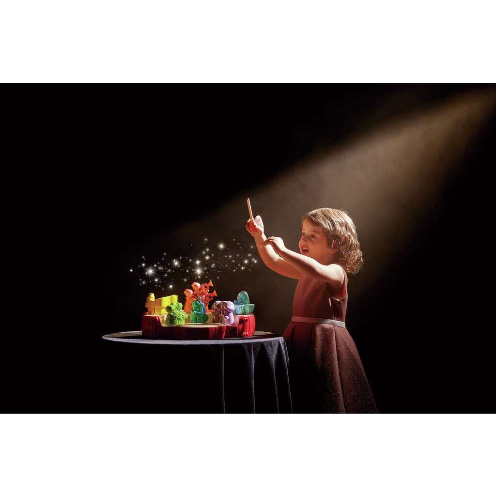 A child stands on a dark stage, holding a wand and pointing at a colorful toy set on a table, with a spotlight illuminating them.