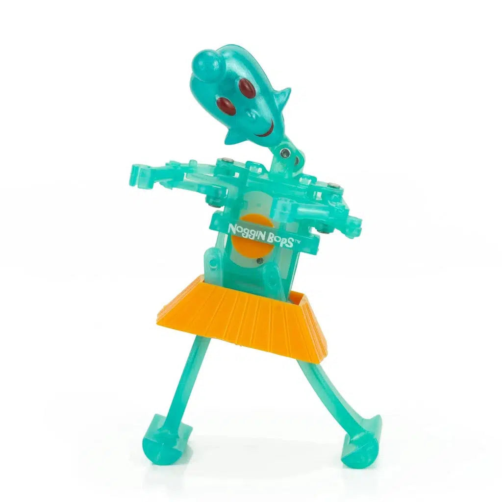 A teal plastic toy figure with a springy head, red eyes, and a yellow skirt performs as a whimsical wind-up dancer. Labeled "Noggin Bops" on the front, this interactive performer is perfect for children aged 3 and up.