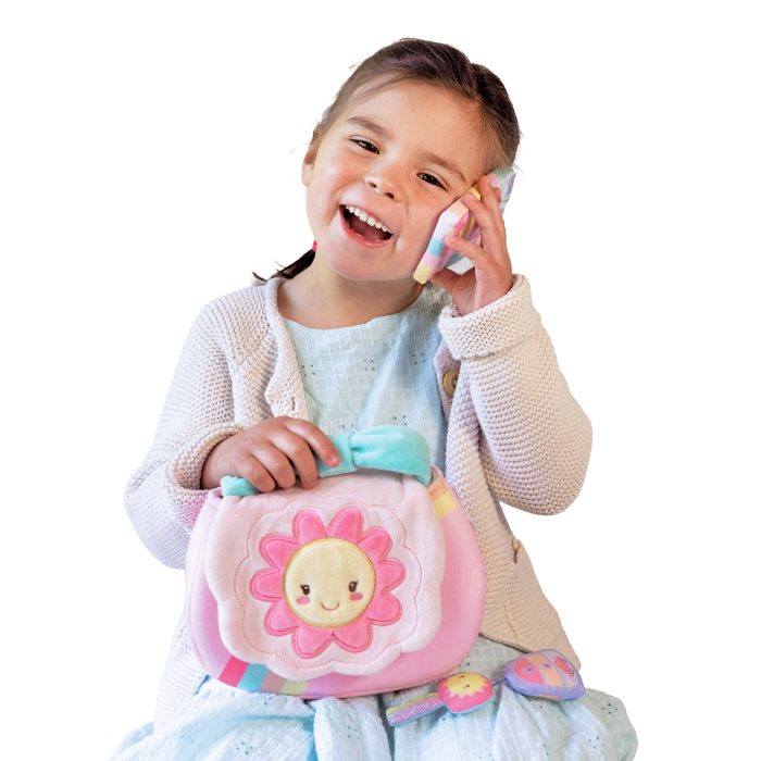 A smiling child holds a pink plush toy, proudly clinging to My First Purse, complete with a charming flower design. Ready for imaginative adventures, this delightful piece is the perfect introduction to plush accessories.