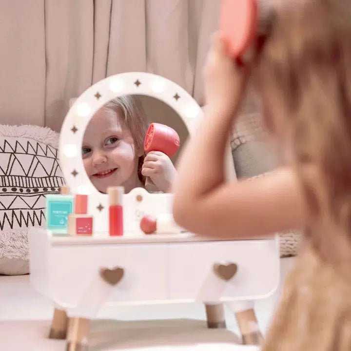 A child engages in imaginative playtime with a light-up dressing table, using a toy hairbrush in front of the illuminated vanity mirror surrounded by realistic accessories and various toy cosmetics.