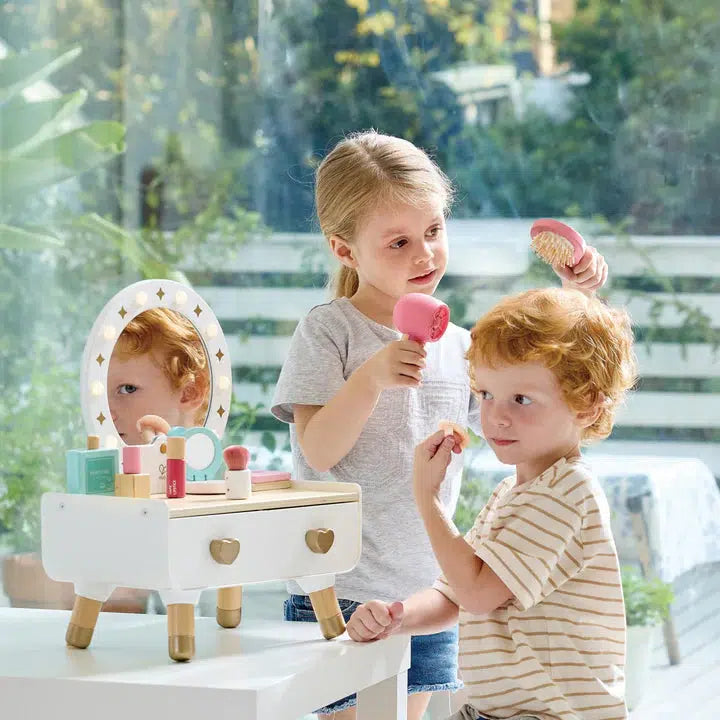 Two children indulge in an immersive makeup experience by a window, surrounded by greenery. One child holds a brush, while the other applies pretend lipstick from their realistic accessories. Their light-up dressing table adds to the enchanting playtime ambiance.