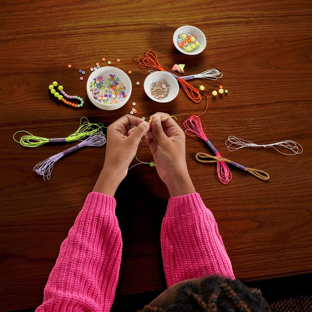 A person in a pink sweater is creating trendy bracelets at a table filled with colorful thread and bowls of vibrant beads.