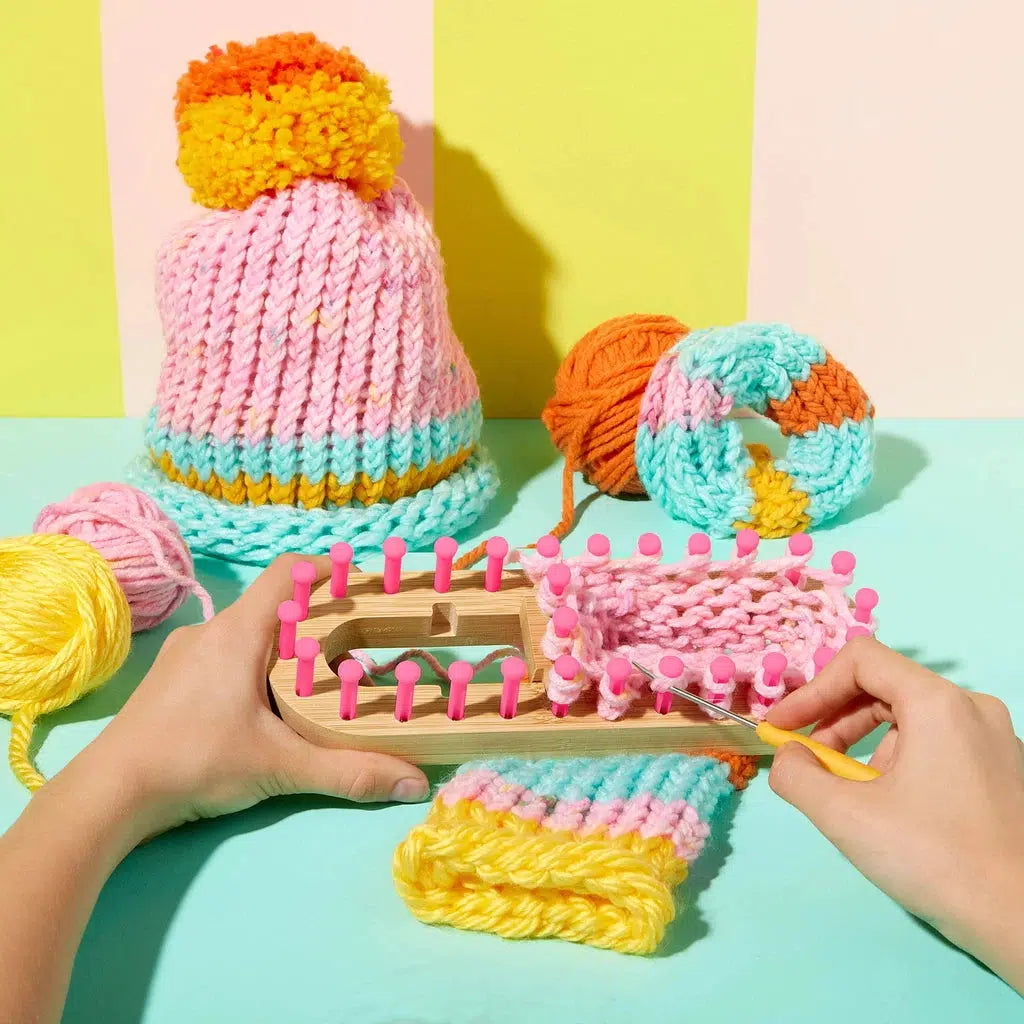 Hands skillfully knitting a colorful hat using a loom, with chunky yarn in pink, yellow, orange, and blue scattered across a vibrant turquoise surface.