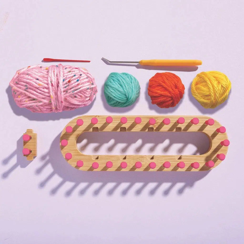 Chunky yarn knitting tools set with a loom, four vibrant yarn balls, a pink hook, and a yellow-handled tool on a light background.