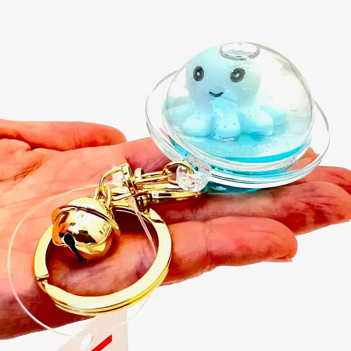 A hand holds an octopus floaty key charm featuring a small blue octopus encased in a clear dome, with a golden bell attached.