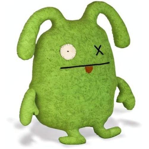 A green plush toy, a classic Ugly Doll, stands upright with floppy ears. It features one button eye and a stitched X for the other, giving it an endearing ox-like charm while a small tongue playfully sticks out.