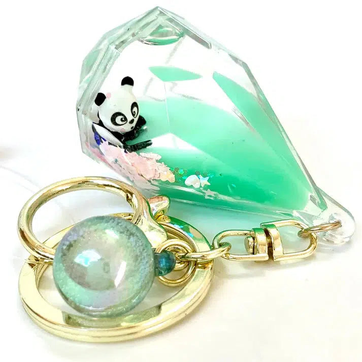 This Diamond Floaty Key Charm features a clear, green gemstone shape holding a tiny panda figure, reminiscent of Panda Boba fun. It comes with a gold ring and a blue-green sphere charm that adds just the right touch of whimsy.