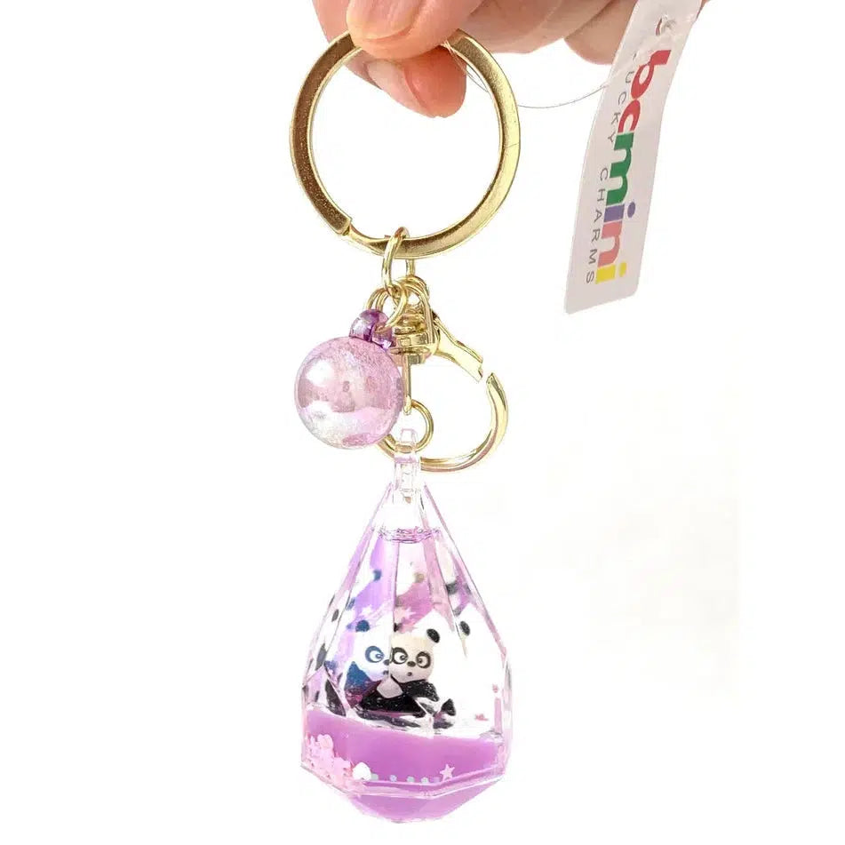 A hand holds a charming keychain with a faceted, clear casing featuring a small panda figure inside. The key charm includes a purple bead and a tag reading "bcmini LUCKY CHARMS," adding whimsy to your keys with its delightful touch of silly animals.