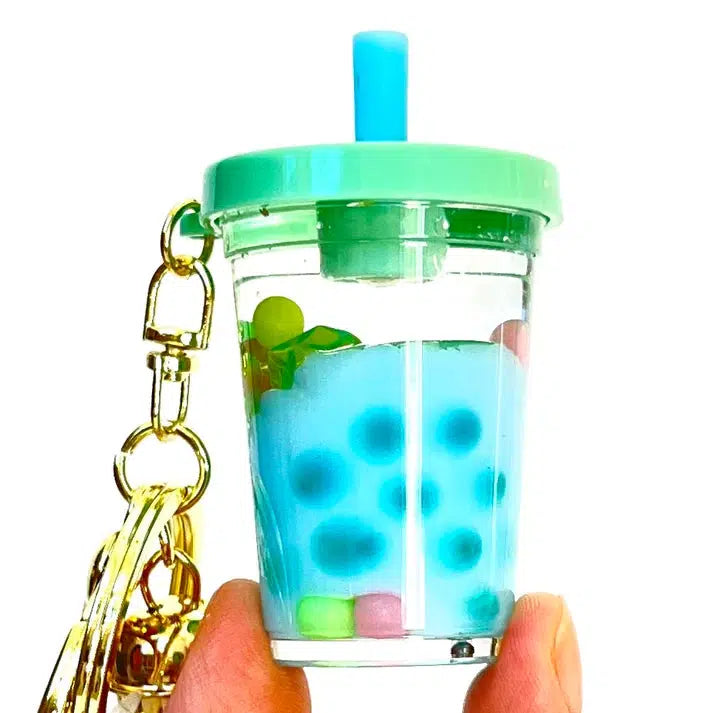 A hand holds a clear miniature cup with a green lid and straw, resembling pastel boba floaty. The cup showcases blue liquid and colorful beads. This whimsical piece doubles as a key charm with a gold keychain attached, celebrating silly animals and foods in style.