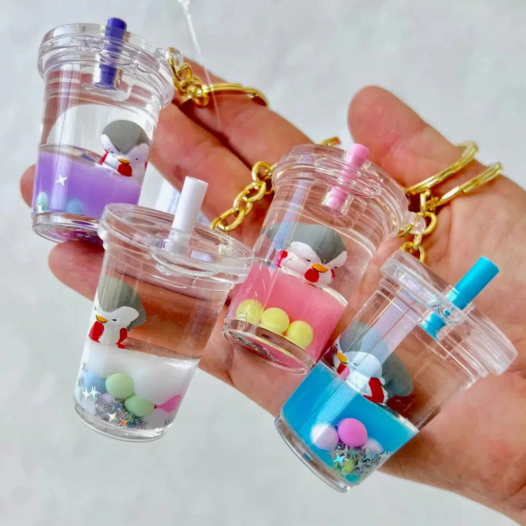 A hand holds four miniature drink keychains adorned with colorful liquids and straws. Each quirky key ring contains a small figure, like a silly animal or penguin floaty charm, accompanied by vibrant beads inside.