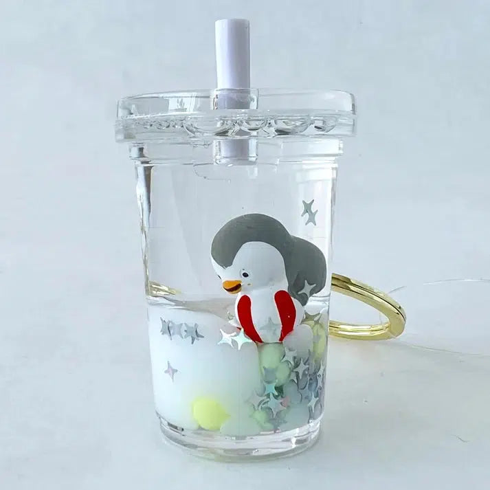 A clear cup features a penguin floaty key charm inside, drifting among colorful beads and star shapes. A straw extends from the lid, and a gold string is attached.