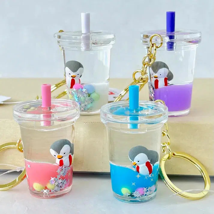 Four clear Penguin Floaty Key Charm cups with toy figurine penguins inside, each filled with different colorful liquids and topped with a matching straw, arranged on a beige surface.