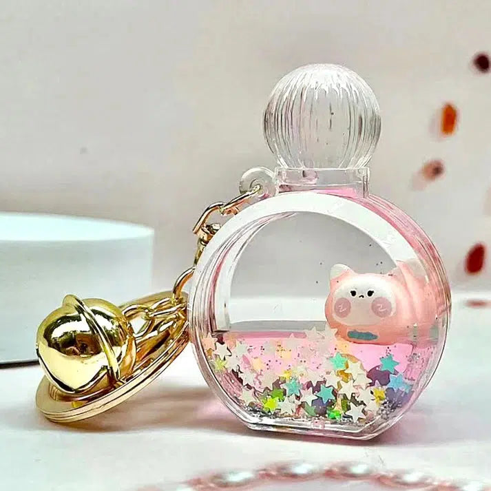 A key ring accessory featuring a round perfume bottle-shaped charm with a small pink cat figure inside, surrounded by colorful stars, attached to a gold planet and bell.