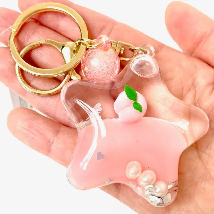 A star-shaped keychain with pink liquid inside, reminiscent of a boba floaty key charm, decorated with a peach, pearls, and small heart details. It’s attached to gold rings and complemented by a pink spherical charm.