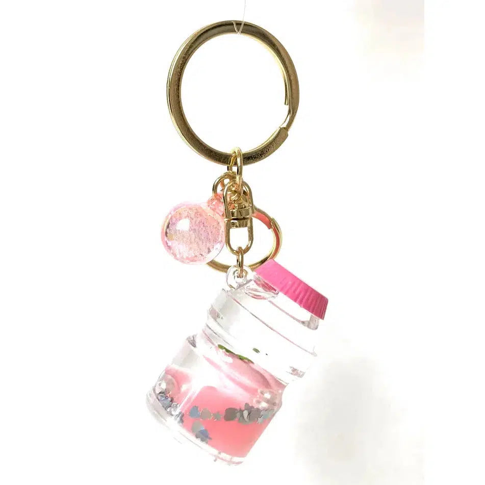 Keychain featuring a small jar charm filled with pink liquid and glitter, complemented by a pink bead and a playful key charm. All elements are elegantly attached to a gold ring, making it perfect for those who love quirky accessories.