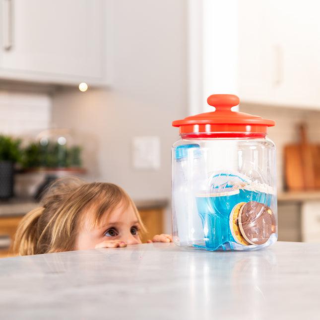 A child peeks over a kitchen counter at a transparent jar filled with cookies, sealed with a red lid.