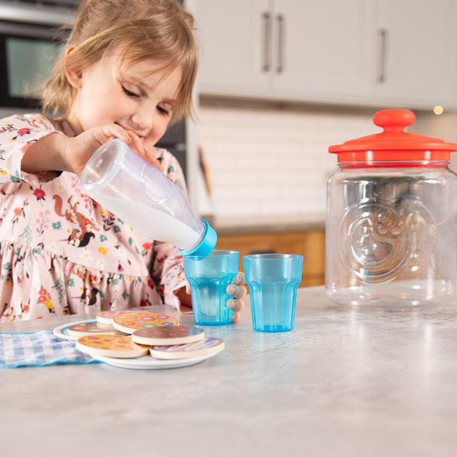 A child pours milk from a bottle into one of two blue glasses on a kitchen counter. A large jar with a red lid and a plate of wooden toy cookies are nearby.