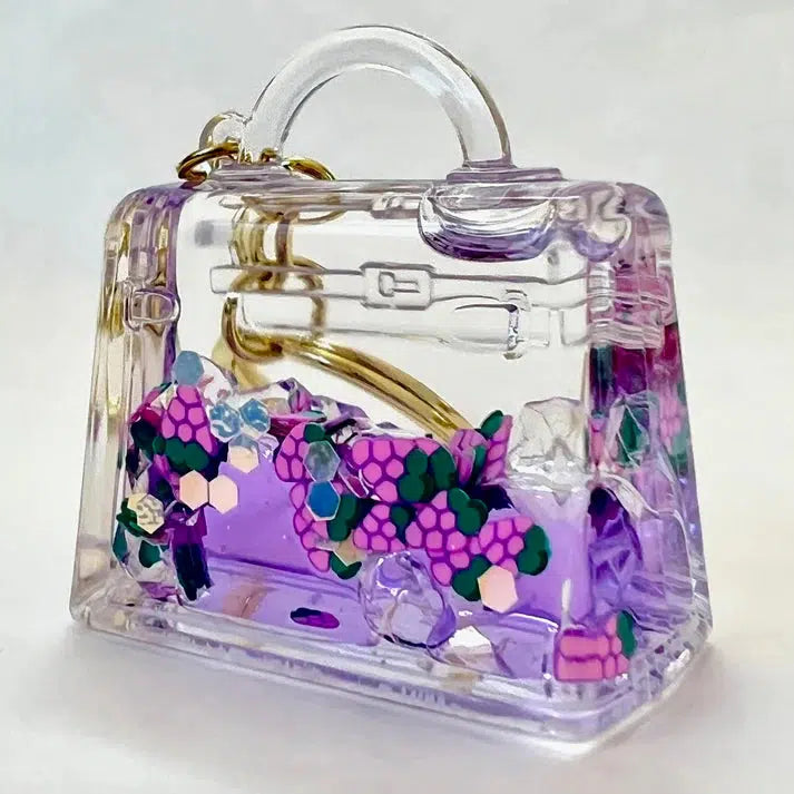 A clear plastic handbag with purple liquid and floating decorative flowers features a top handle, a gold-tone clasp, and a whimsical key charm adding subtle elegance.