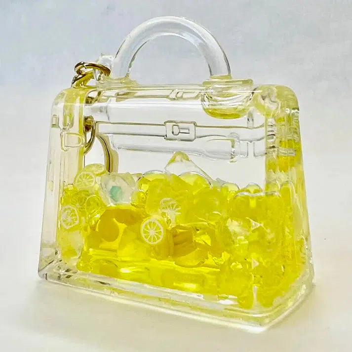 A clear handbag-shaped container filled with yellow candy pieces resembling citrus slices, complemented by a playful key charm, set against a plain background.