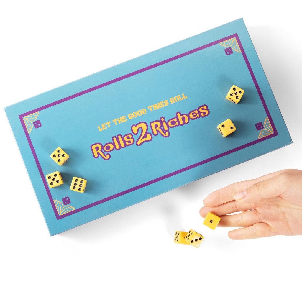 A hand rolling yellow dice onto a blue board game box labeled "Rolls 2 Riches" with the phrase "Let the Good Times Roll".