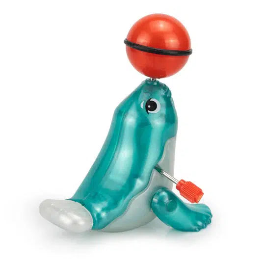 A blue and gray plastic performing seal toy with large eyes balances a red ball on its nose, bringing a touch of circus-style performance to playtime. Equipped with a red wind-up key on its side, this interactive toy promises hours of fun.