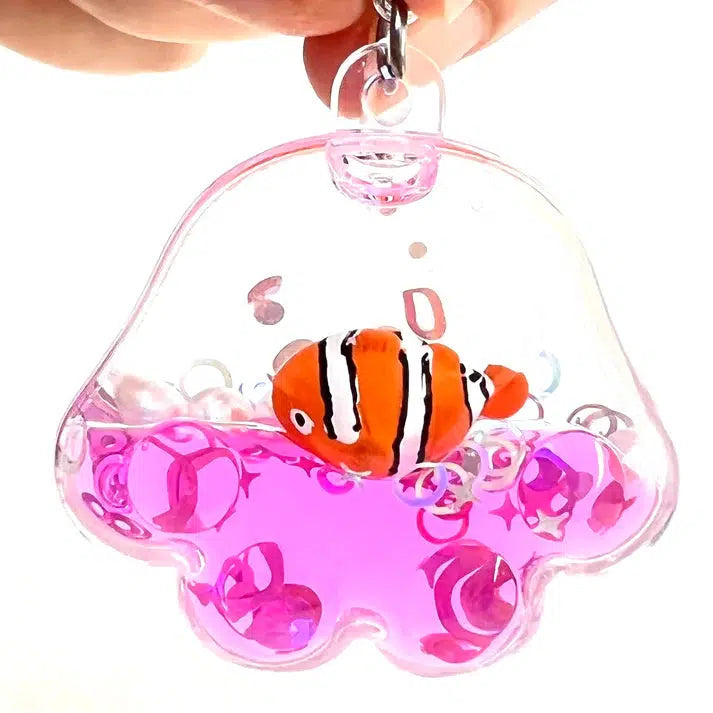 A hand holds a transparent, jellyfish-shaped key charm containing pink liquid and a small toy clownfish inside, making it a delightful sealife-inspired bag accessory.