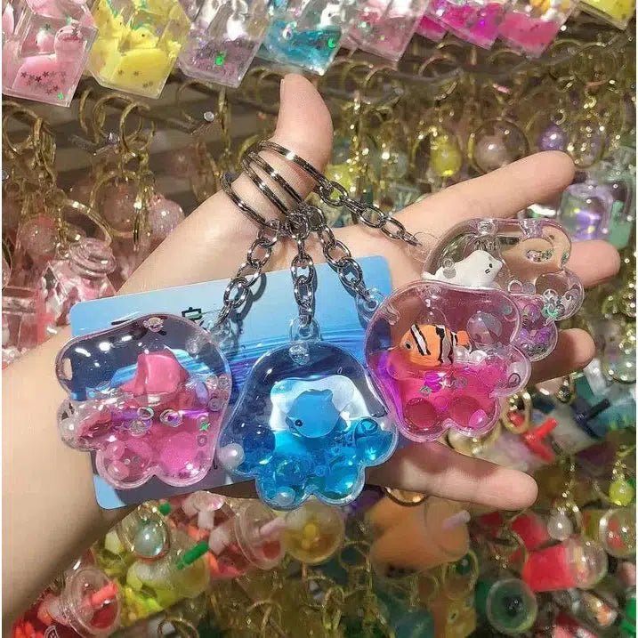 A hand holds colorful, transparent keychains shaped like various sea creatures, with small decorations inside each one, reminiscent of the SEALIFE JELLYFISH FLOATY collection. More assorted designs are blurred in the background.