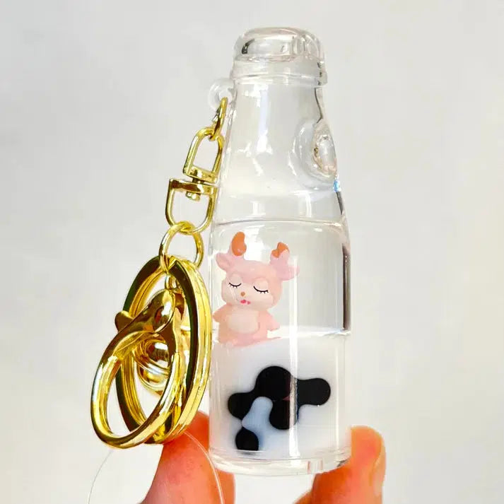 A hand holds a keychain shaped like a milk bottle with a small pink deer figure and black cow spots inside, showcasing one of the many assorted designs. This charming key charm captures the whimsy of silly animals, adding a playful touch to any collection.