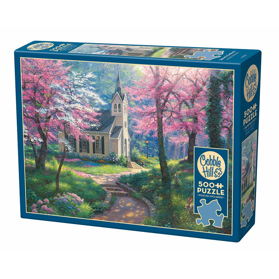 A 500-piece jigsaw puzzle box from Cobble Hill featuring an image of a quaint cottage surrounded by blooming pink and purple trees in a serene, wooded area.