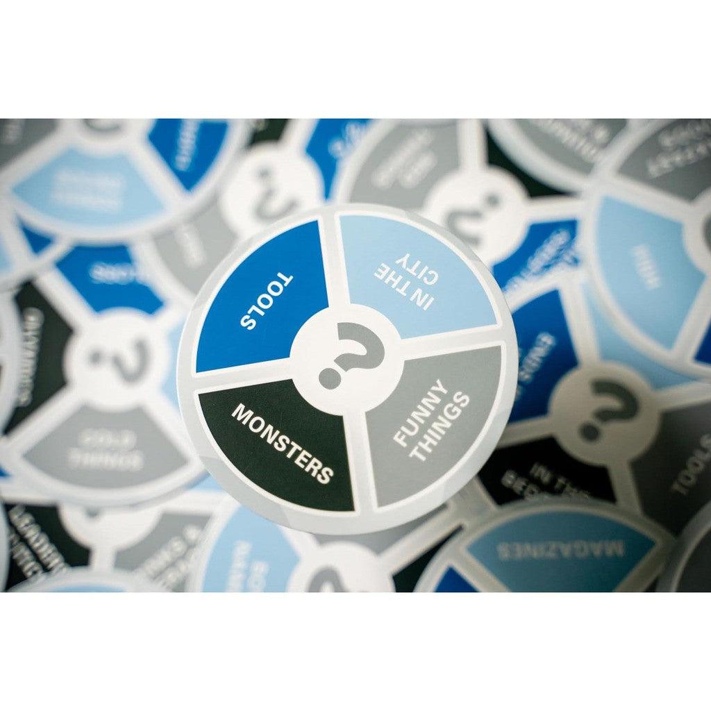 A close-up of circular cards in portable packaging with labeled sections: Tools, Monsters, In the City, and Funny Things. Designed for a family card game, the predominantly blue, gray, and white cards promise fast-paced fun for everyone.