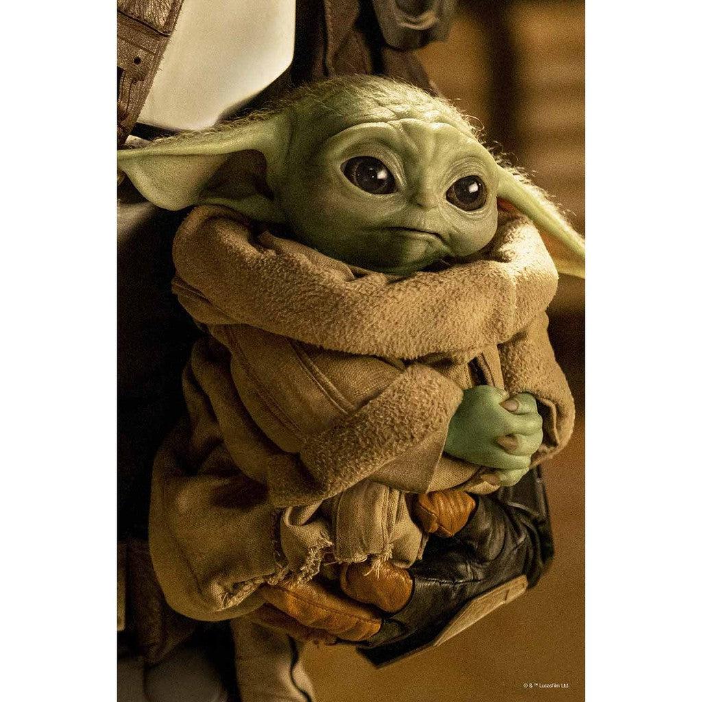 Grogu, a small, green creature with large eyes and long ears, wearing a brown robe, is cradled by the Mandalorian -someone in dark armor and gloves—the iconic duo from the Mandalorian Star Wars saga.