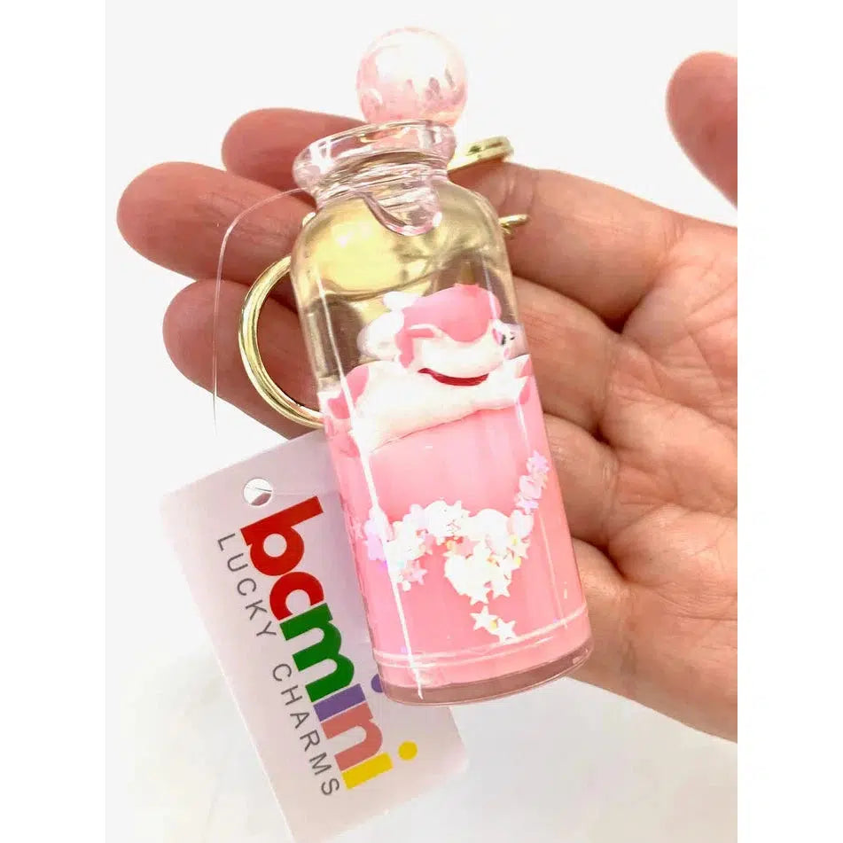 A hand holds a small glass bottle with a pink, white, and red charm inside. The attached tag reads "bamini lucky charms," and peeking out is a miniature key charm shaped like unicorn love—a whimsical touch that captures the magic within.