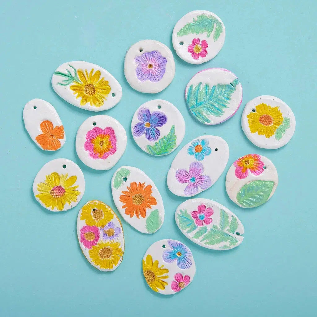 Several white clay pendants featuring colorful embossed flowers and leaves are artfully arranged on a light blue background, showcasing the beauty of ceramic jewelry enhanced with intricate botanical stamps. 