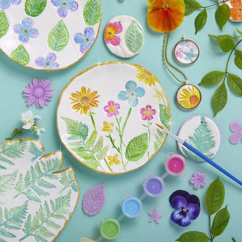 Decorative plates and circles with floral and leaf designs, surrounded by real flowers, leaves, and a paintbrush with paint pots on a teal background. The scene is reminiscent of crafting ceramic jewelry pendants using botanical stamps.