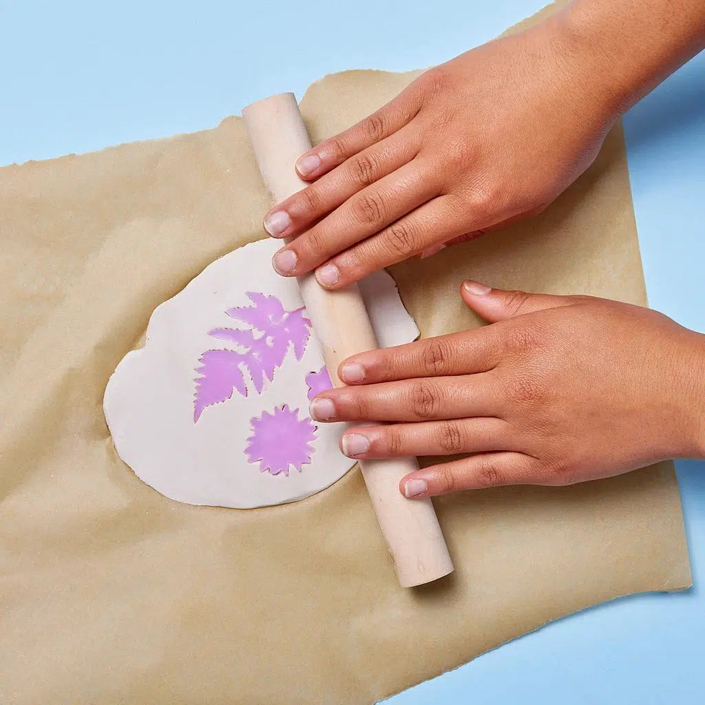 Hands use a rolling pin to press a purple leaf and flower pattern into air dry clay, creating intricate designs on parchment paper perfect for crafting ceramic jewelry.